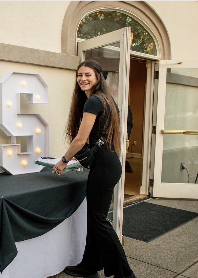 Serena smiling beside an event table, holding a clipboard and wearing her event headset.